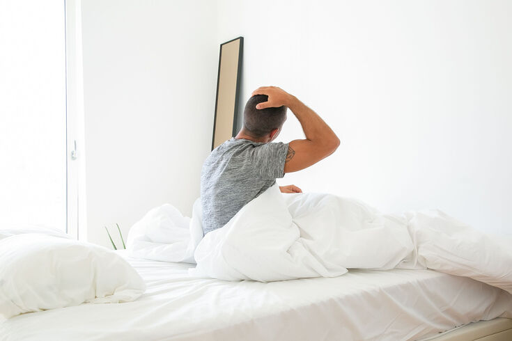 Man sitting on the edge of a rumpled bed with his hand behind his head
