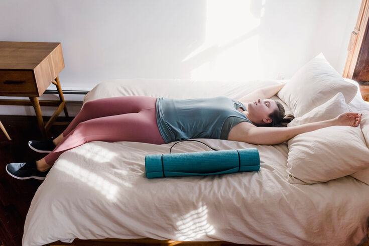 Woman in workout clothes lying back across a bed beside a rolled yoga mat