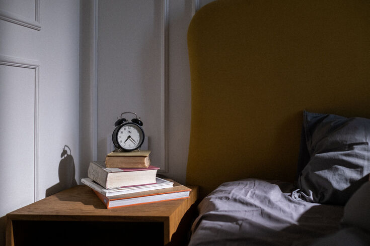 Alarm clock on a stack of books on a nightstand beside a bed with grey bedding